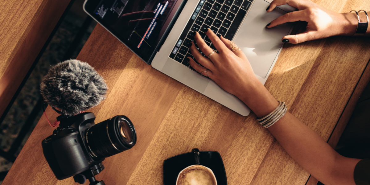 Opengeklapte laptop met twee vrouwenarmen die typen op het toetsenbord. Naast de laptop ligt een spiegelreflexcamera en er staat een kop koffie.
