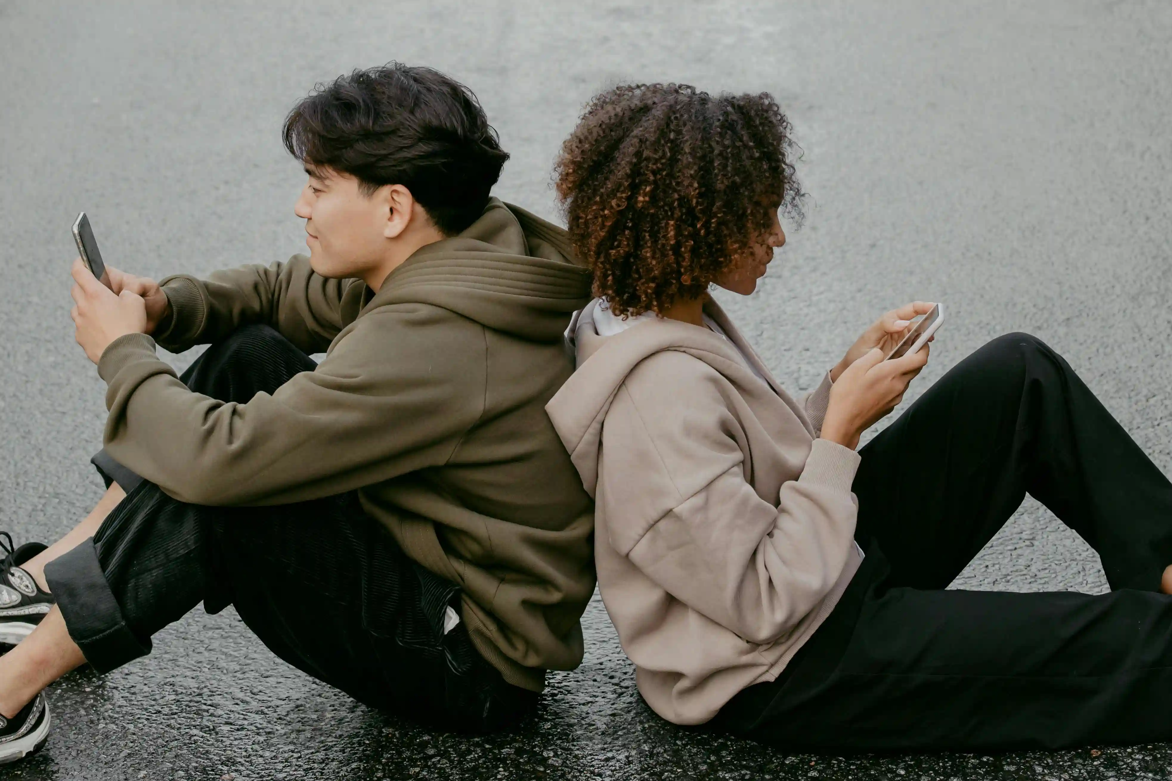 Foto van twee jongvolwassenen die rug-aan-rug op hun mobiele telefoon aan het kijken zijn.
