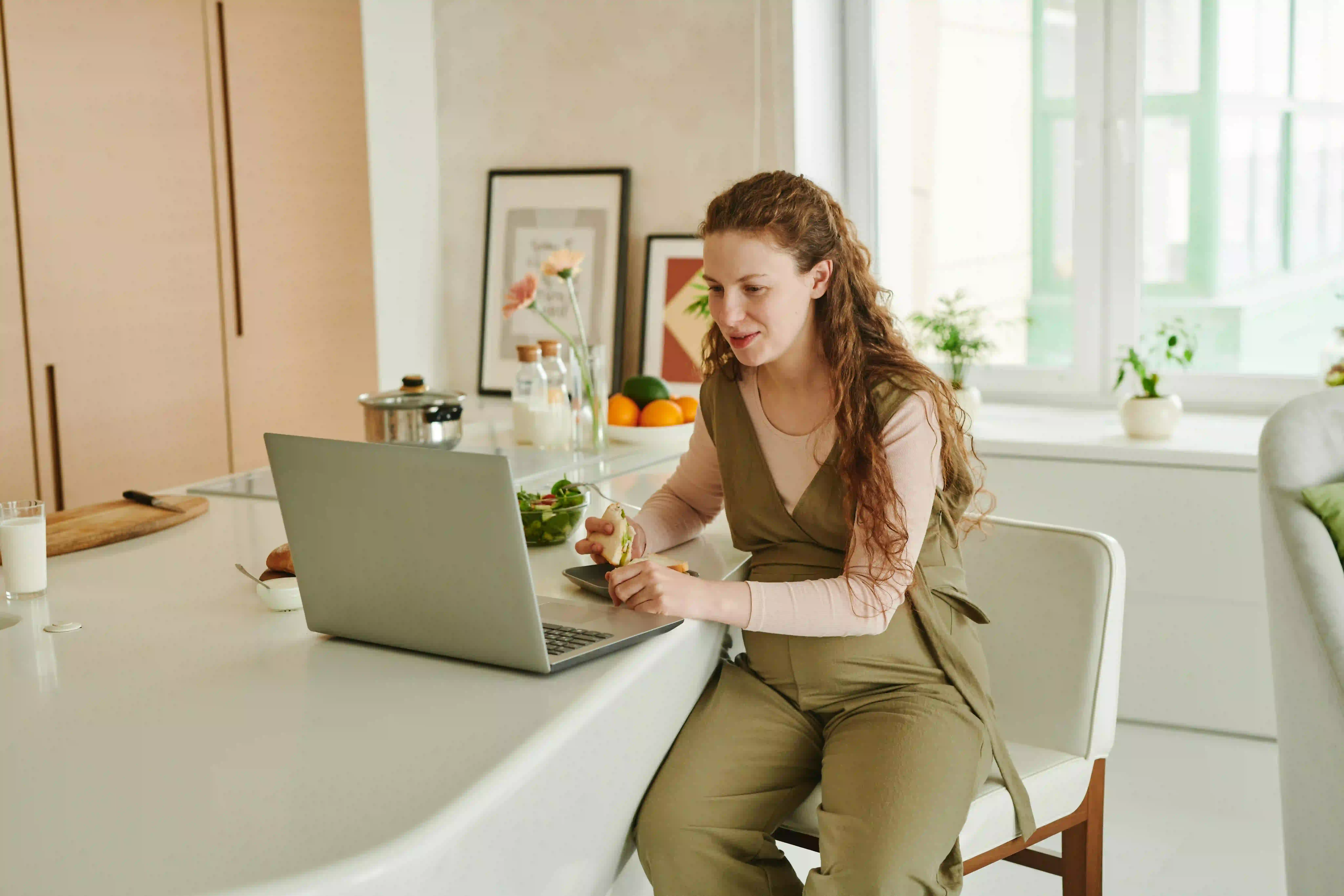 Foto van een zwangere vrouw die aan een keukentafel eet met haar laptop voor haar.