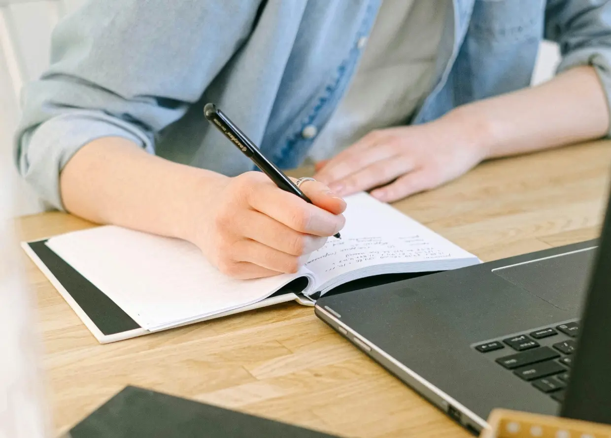 Persoon die notities neemt in een notitieboek met een opengeklapte laptop